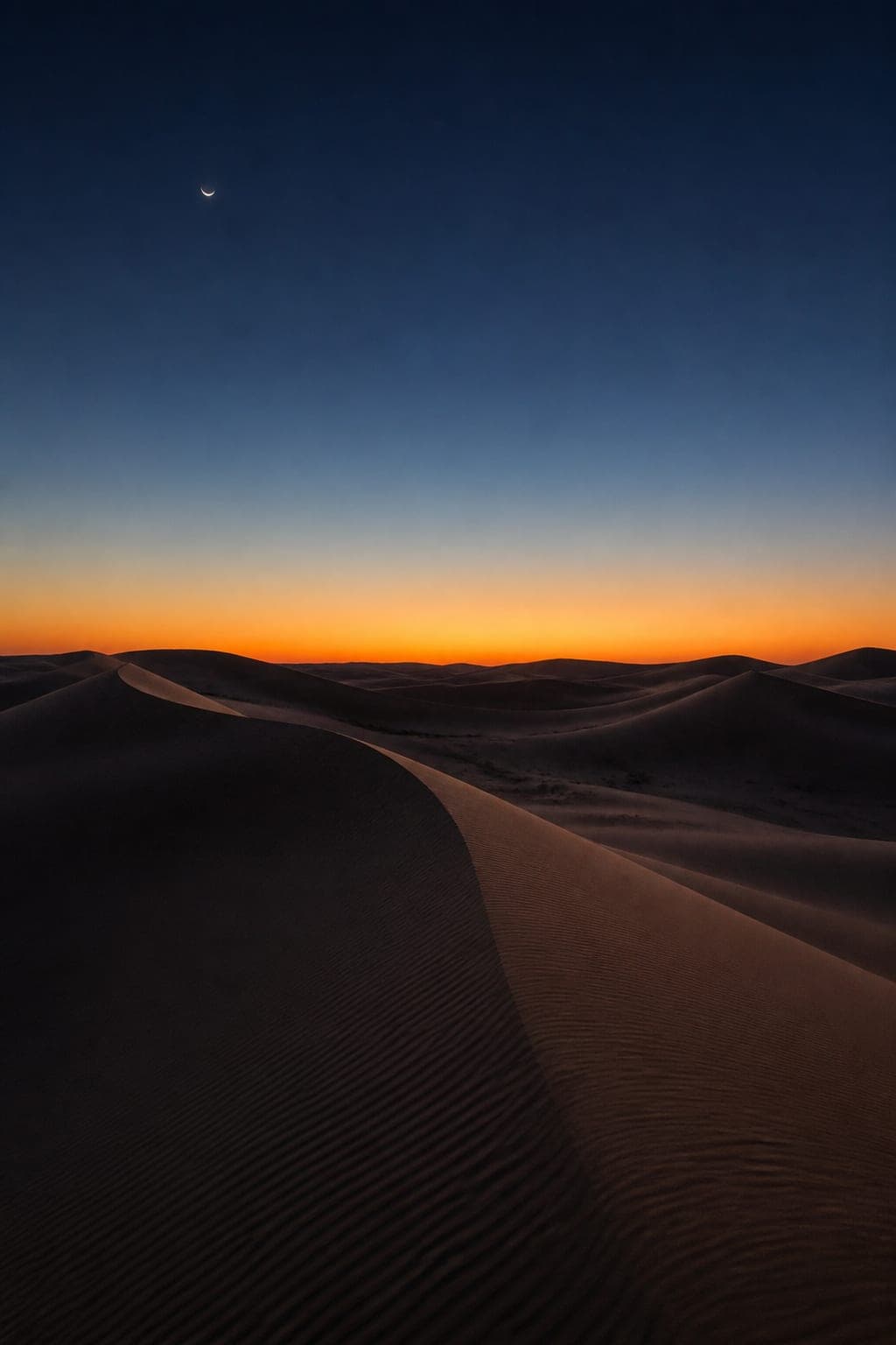 Desert dunes beneath a crescent moon at blue hour