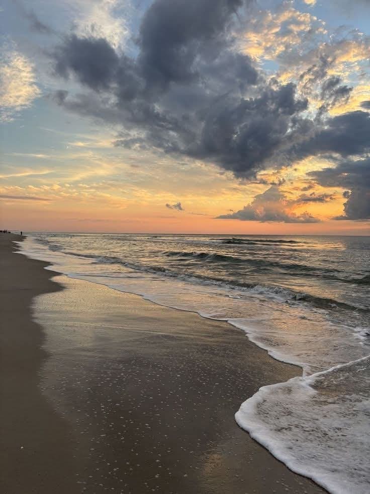 A sandy beach at sunset with white sea foam on wet sand and dark clouds over an orange horizon. Sunset photograph.