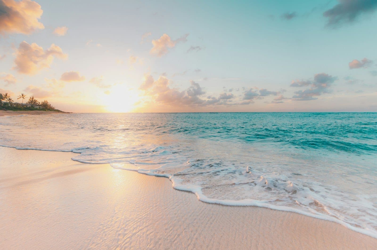 A sandy beach and turquoise water under a peach and pink sky, with palms along the far shore